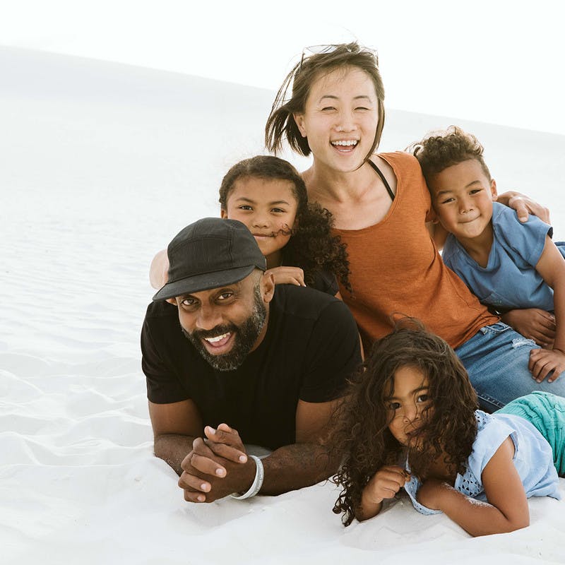 The Register Family, man and woman with three kids, lay in white sand and smile at the camera.