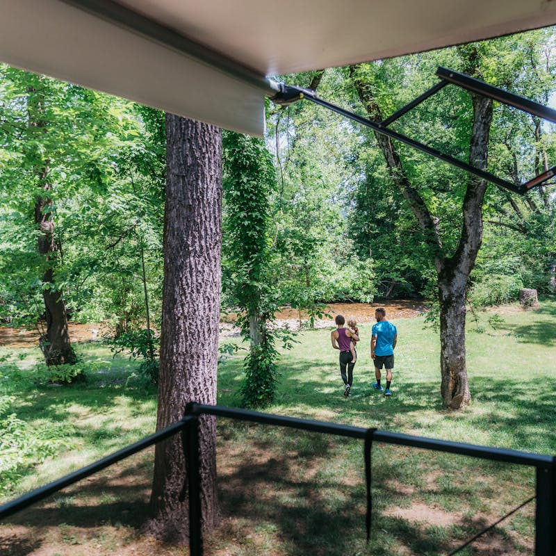 A young mom and dad walk their toddler daughter across a grassy lawn toward a stream.