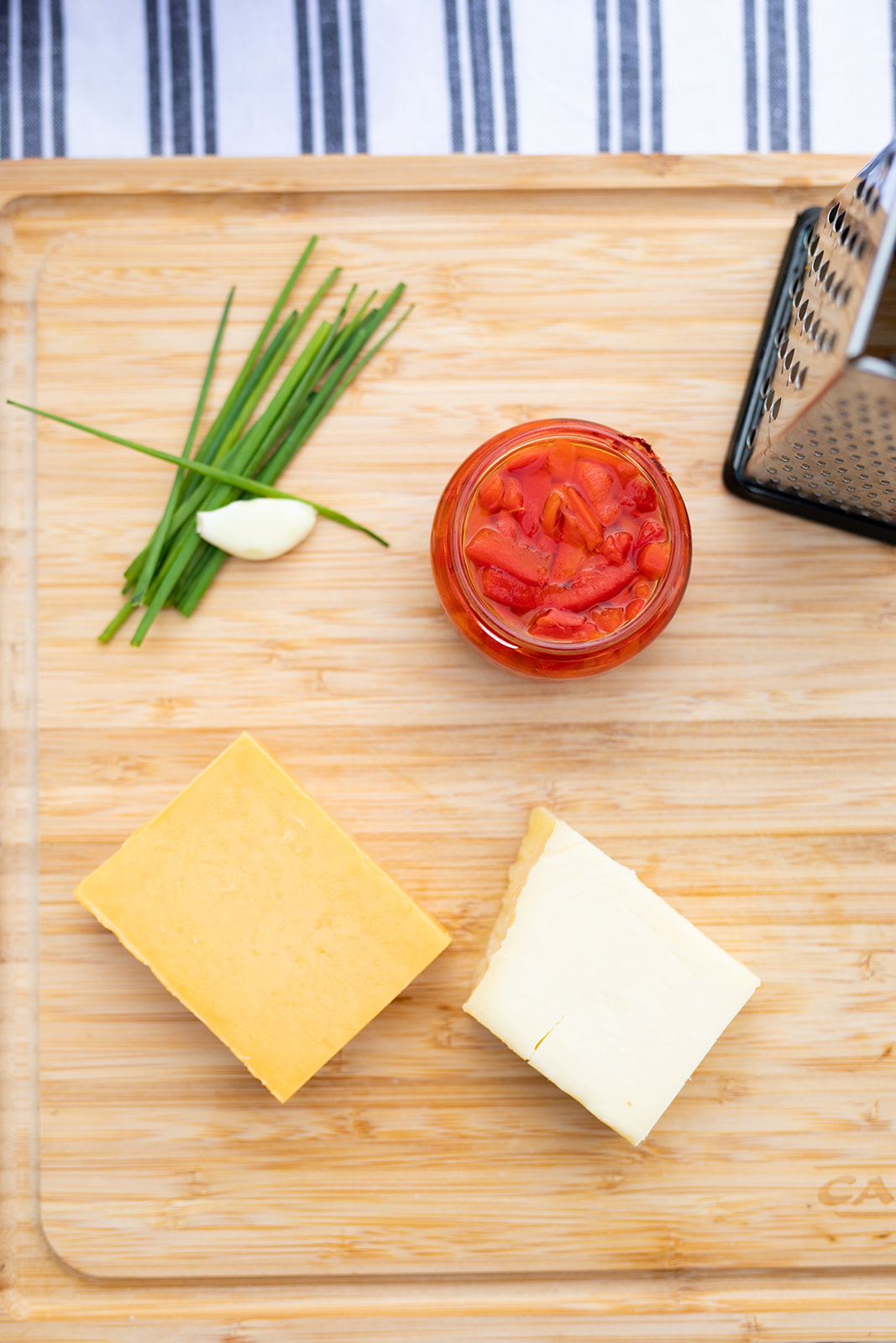A cutting board with chives, garlic, cheese and a jar of pimentos on it.