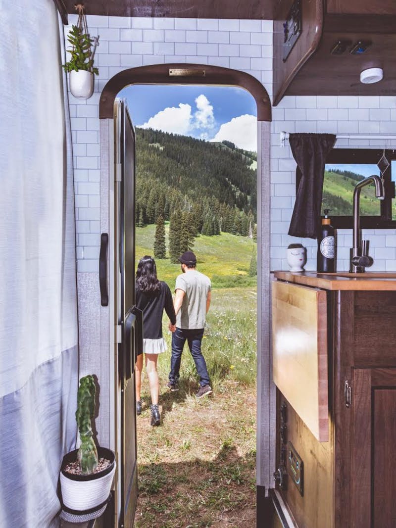 A man and woman holding hands outside their RV door walking towards the mountains filled with trees.