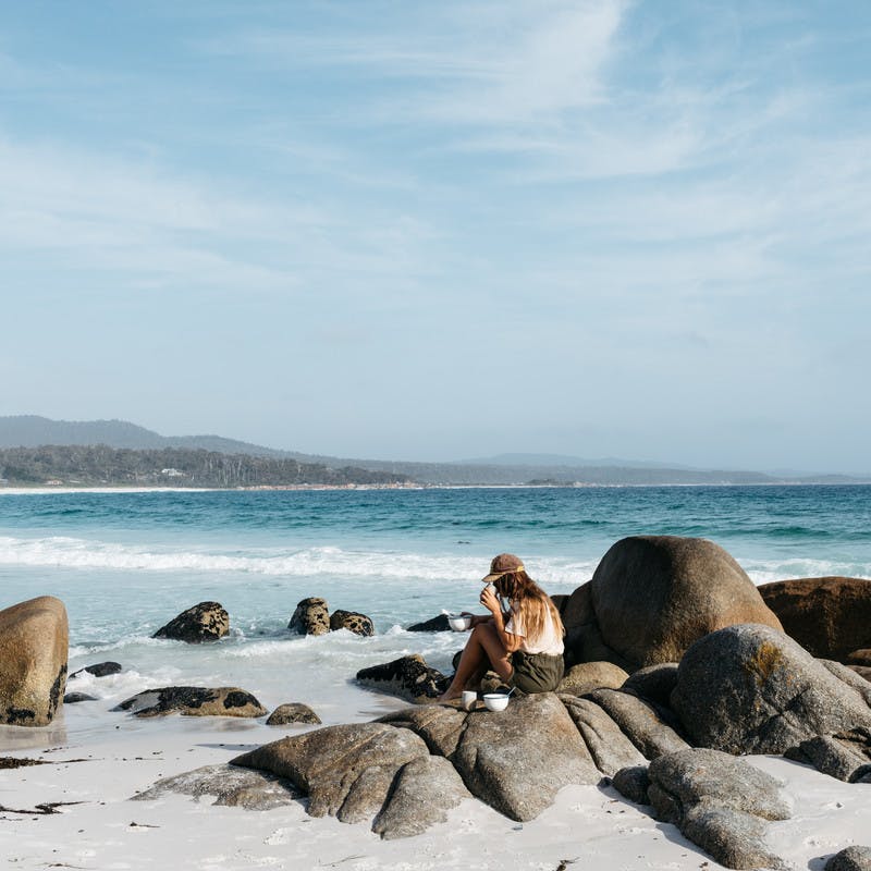 Sarah Glover drinking coffee and eating oatmeal while she sits on a smooth boulder on the beach with the ocean in the background.