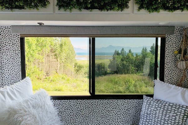 A view from the inside of an RV with comfortable pillows, polka dot wallpaper and a large window looking out to green trees and mountains in Homer, Alaska.