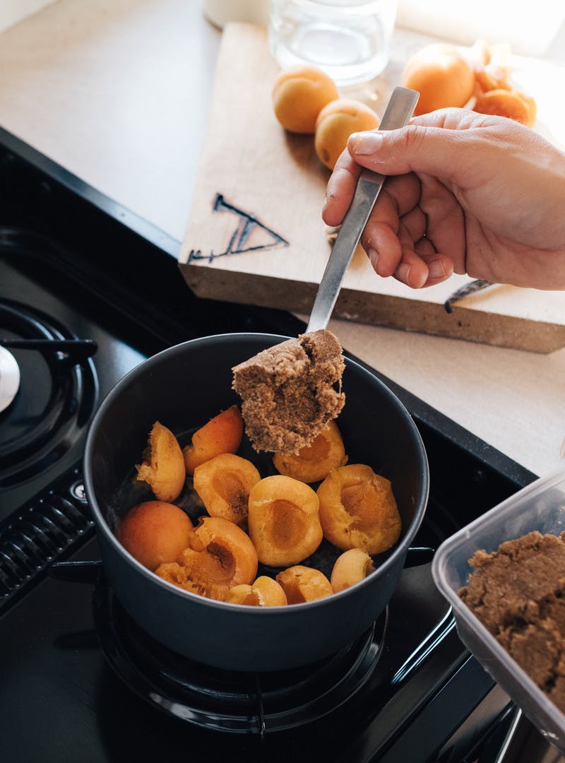 Rosy-orange apricots cut in half inside a small stock pot, with a hand adding a spoonful of brown sugar to the pot.