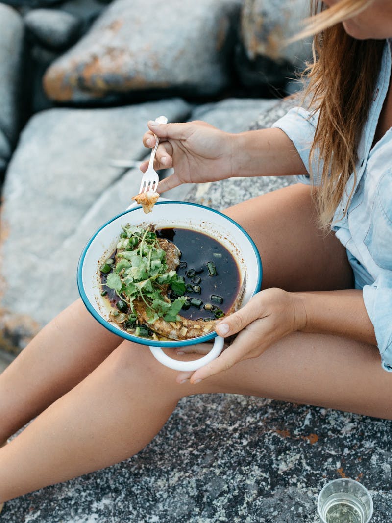 Close up of woman's legs, eating fish in brown broth with cilantro and a fork.