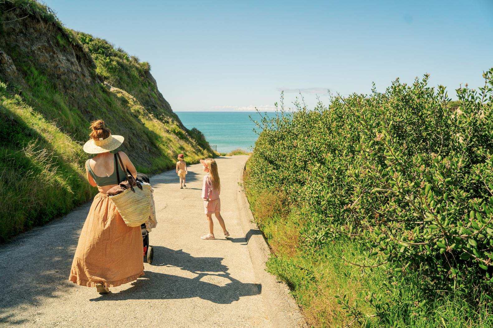 Mom in orange skirt pushes stroller down paved road with two kids and ocean in background