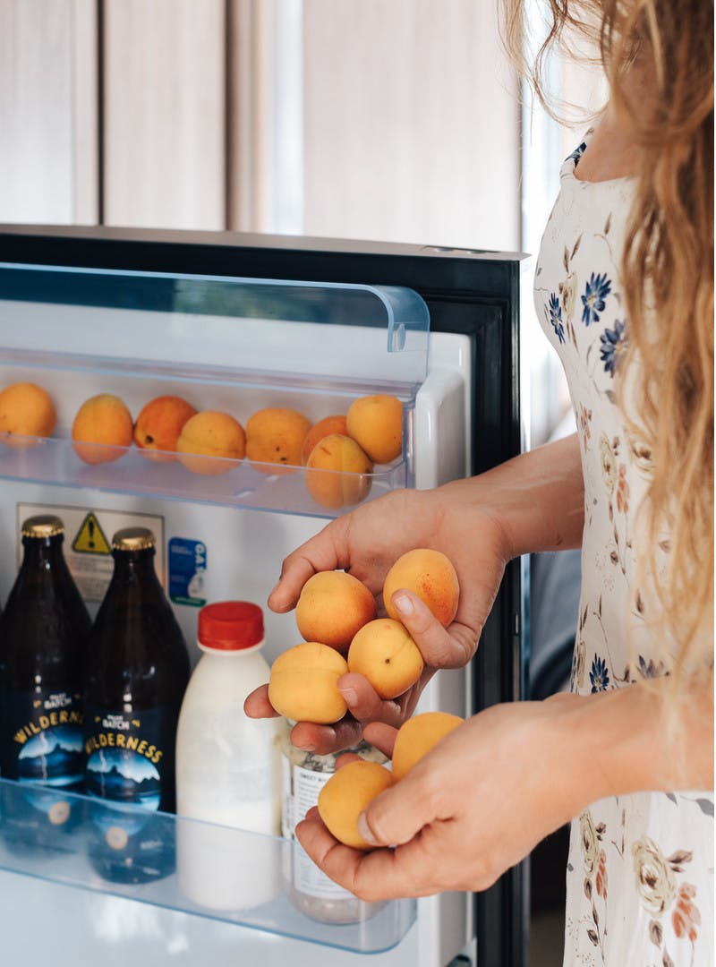 A close shot of a woman standing with the refrigerator door open. Her hands and the door are filled with fresh apricots.