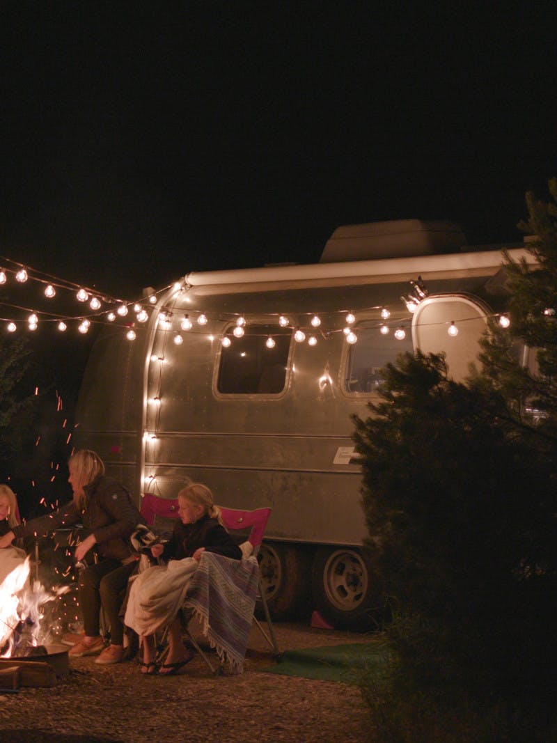Family sitting around fire in front of Airstream, hung lights. 