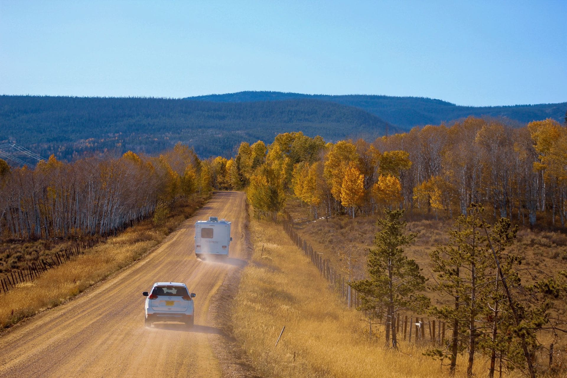 White car and Class C RV on dusty dirt road heading into forest of yellow trees