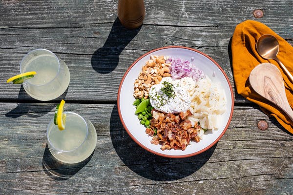 White bowl on picnic table filled with peas, bacon, red onion, water chestnuts, cashews, and sour cream, with two glasses of lemonade.