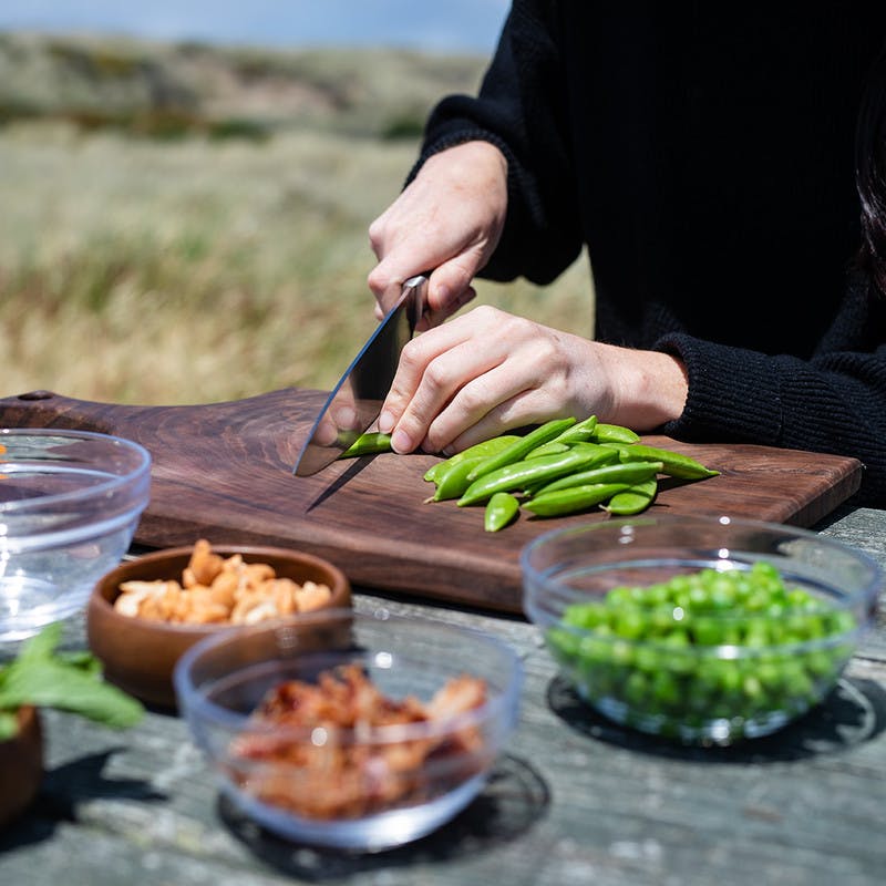 Picnic table with small glass bowls, and a woman slicing snap peas on a cutting board.