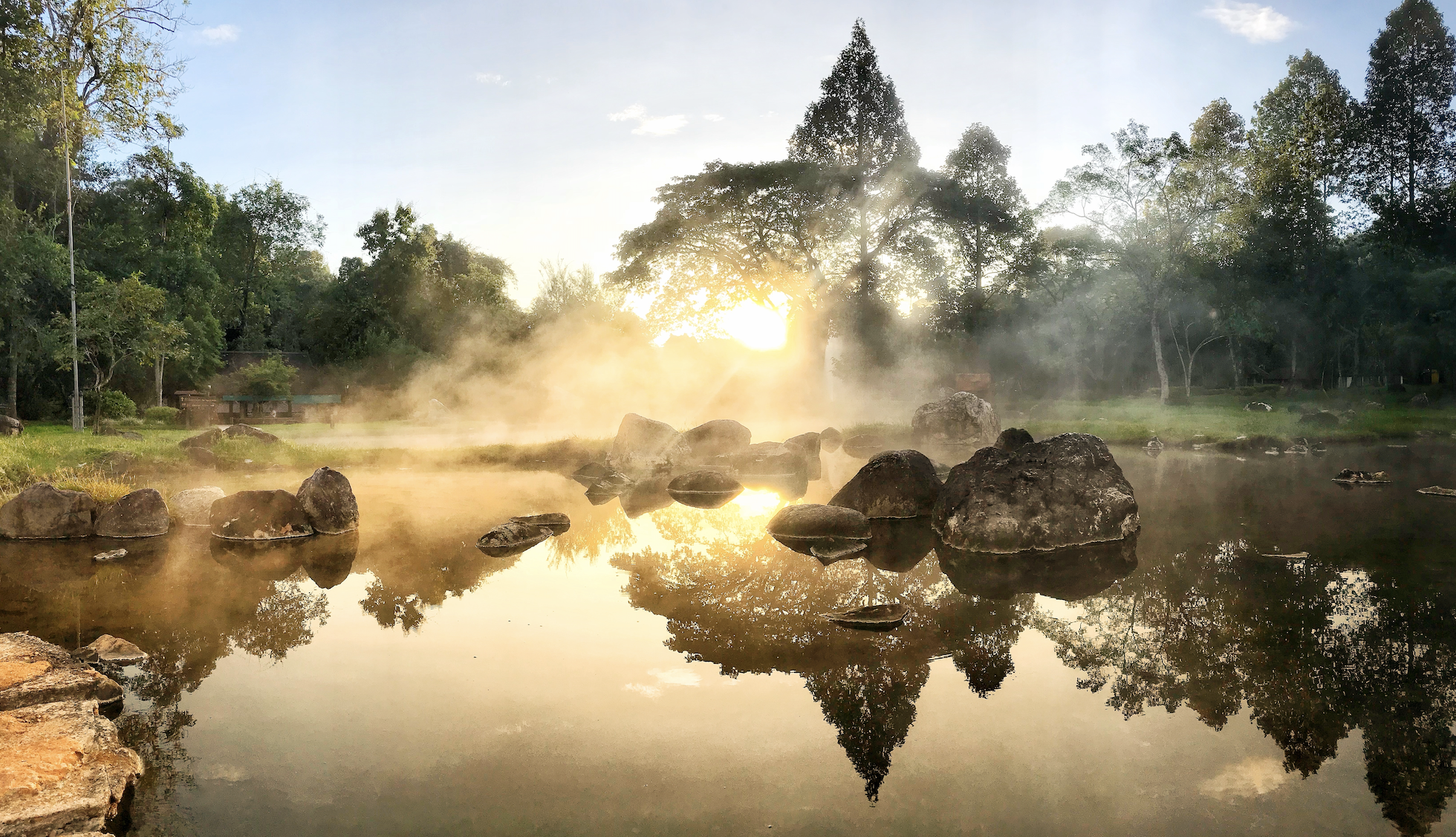 Golden sunrise reflects on a glassy lake with steam rising and tall trees in the background