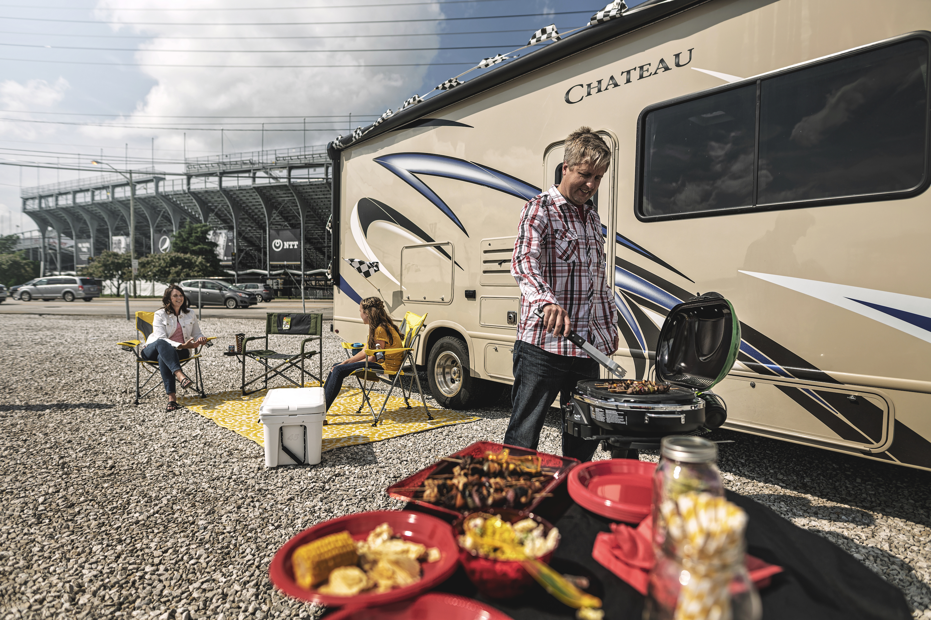 A man grilling near at Class C RV parked outside of a stadium. 