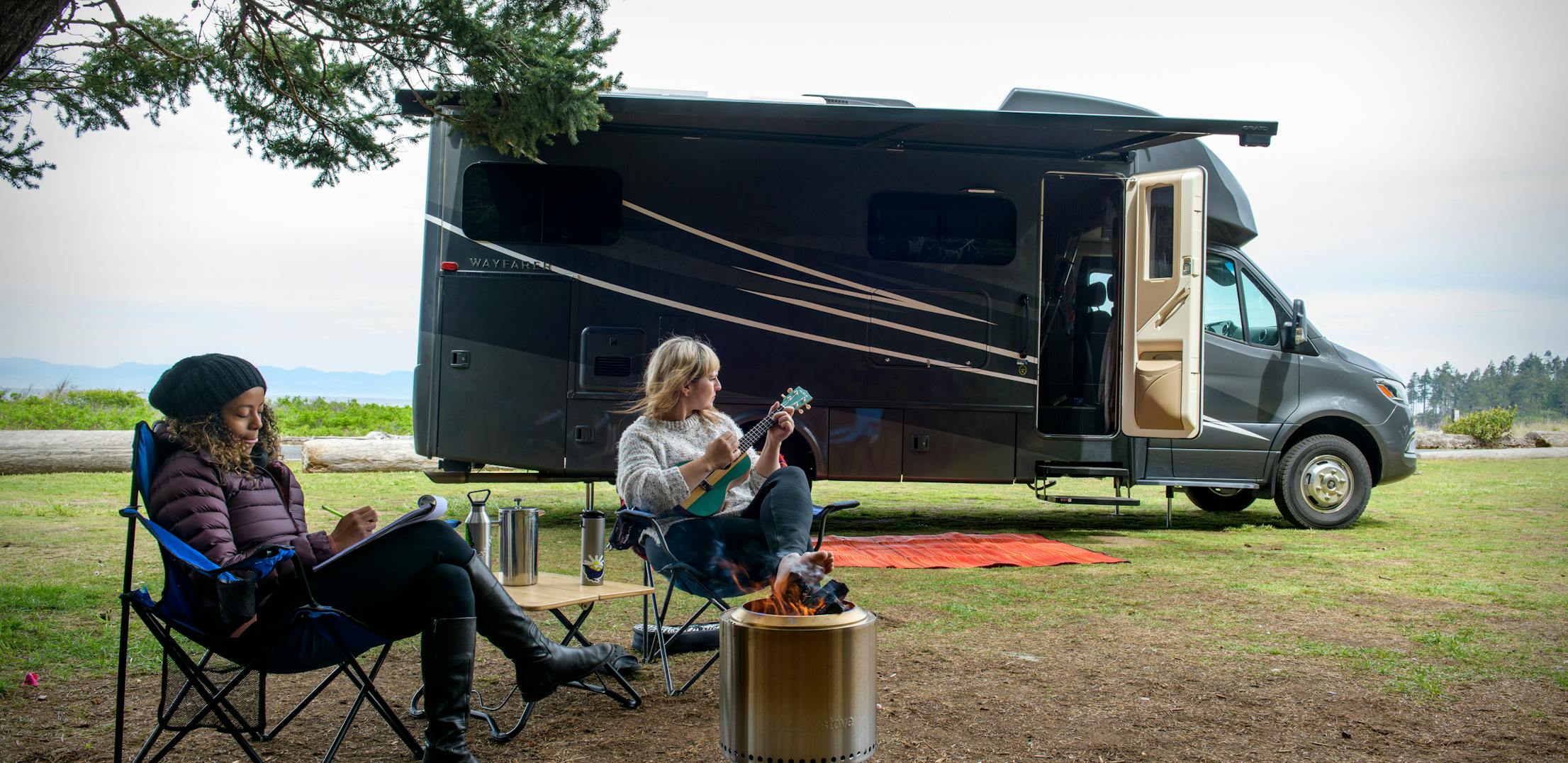 Two women sit around a campfire outside of a Tiffin Wayfarer.