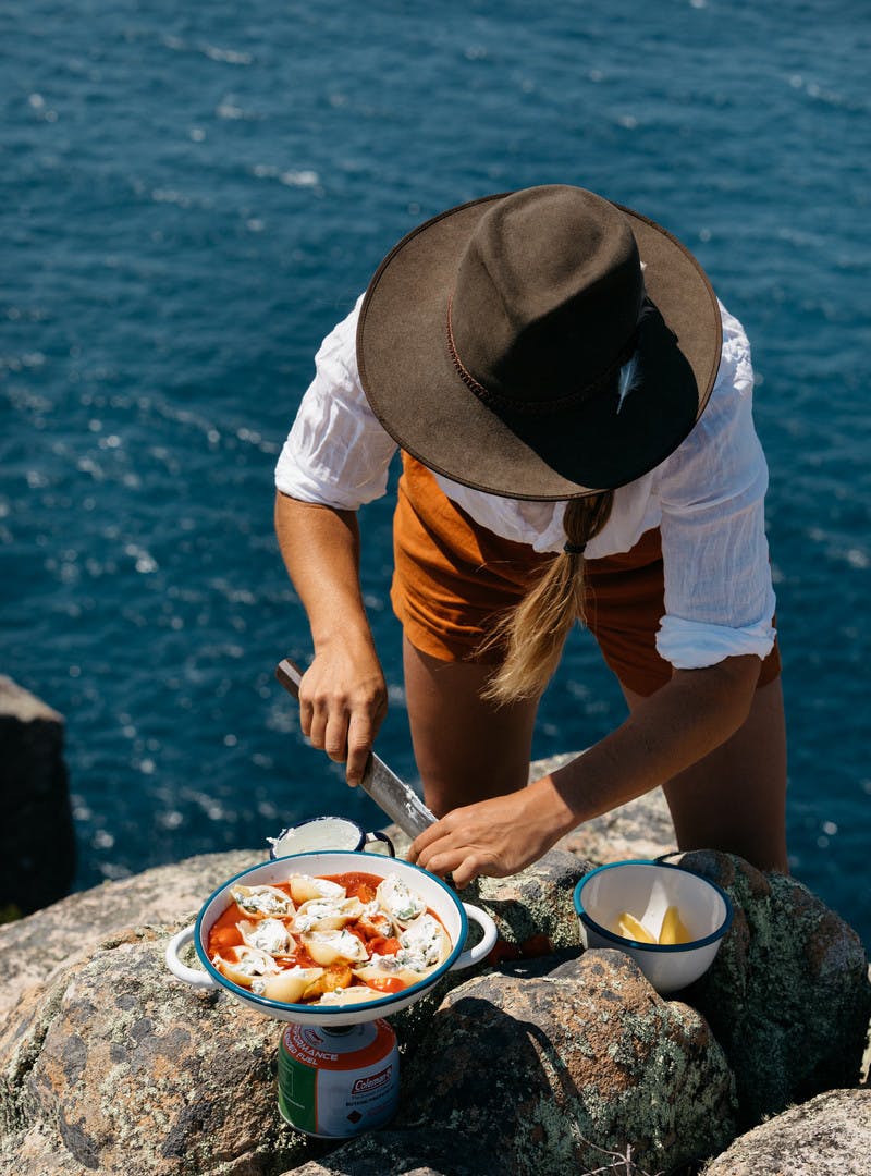 Sarah Glover with a large knife cooking pasta shells on a camping stove over the water.