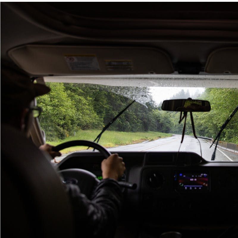 View out front windshield of motorhome onto road and forest with windshield wipers.