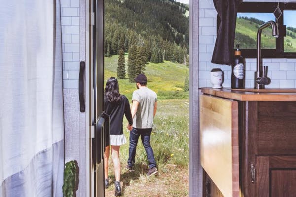 A man and woman holding hands outside their RV door walking towards the mountains filled with trees.