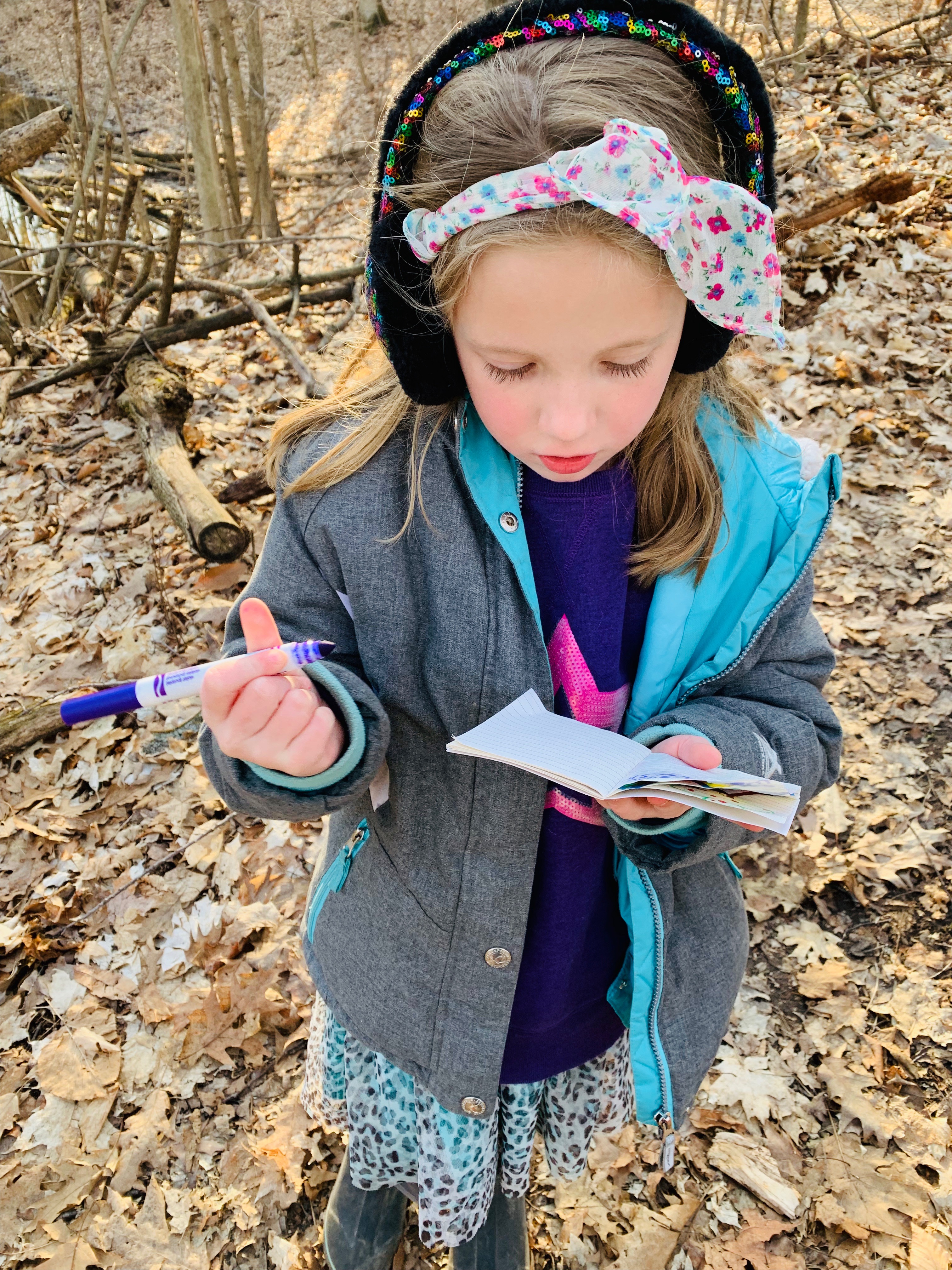 Young girl with headband and earmuffs holds purple marker and notepad in the forest