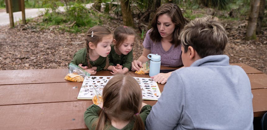 Two moms and three triplets looking at animals in a book while sitting outside at a picnic table.
