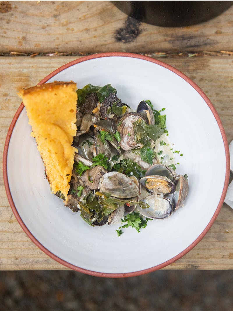 Bowl of cooked clams with parsley on a white plate, a golden slice of cornbread, and gold utensils on a white napkin.