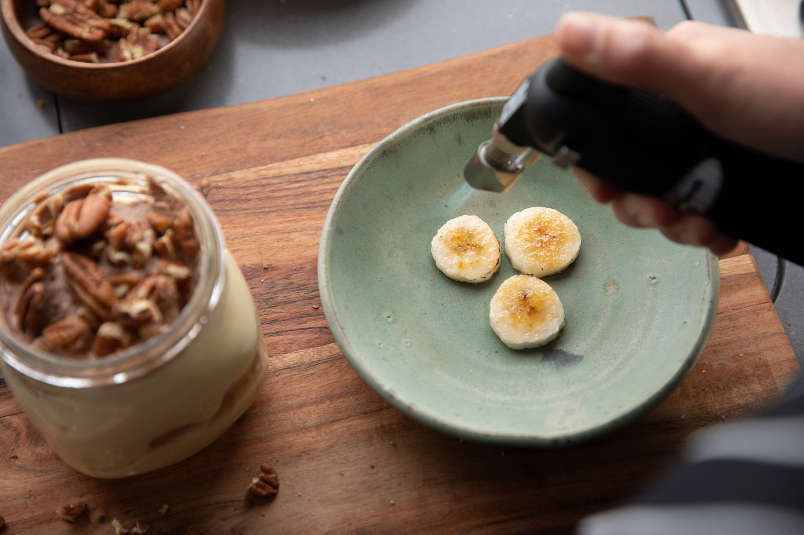 A hand using a kitchen blowtorch to caramelize the tops of banana slices.