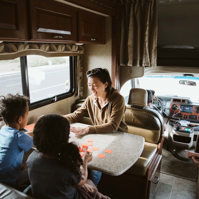 Inside RV, woman in brown jacket sits at table with kids and plays a card game