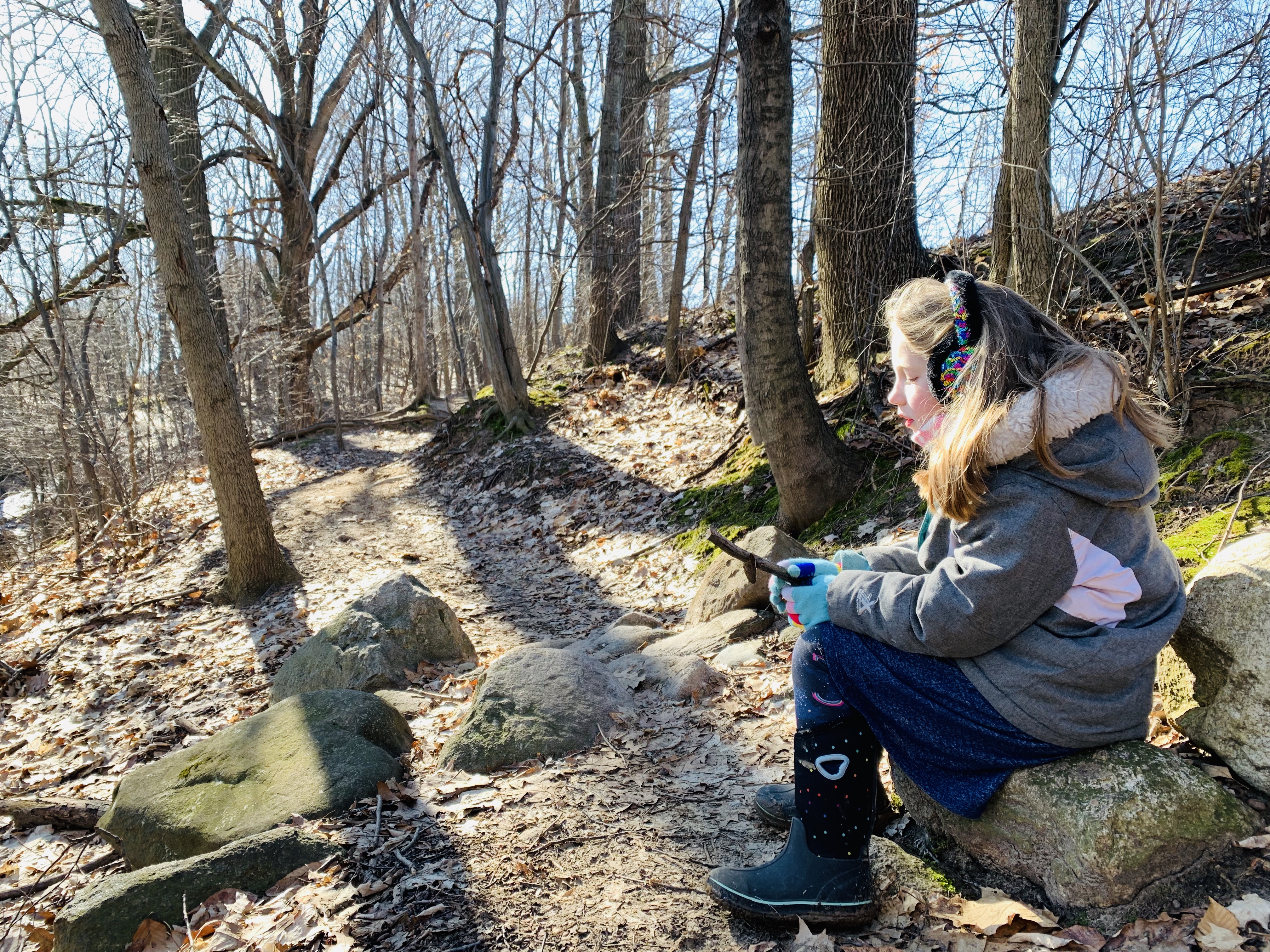 Young girl wearing snow jacket and earmuffs sits on a long and admires a stick