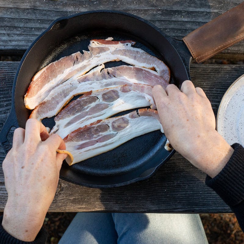 Woman lays raw bacon on pan to prepare to cook.