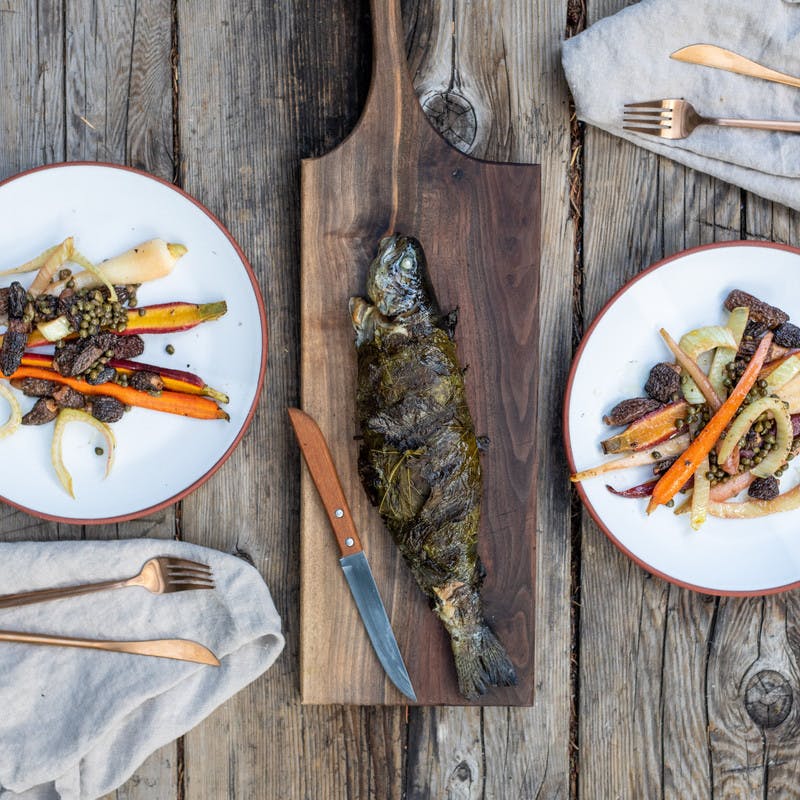 Photo from above of two white plates filled with cooked carrots, morels and fennel, and a full cooked trout wrapped in grape leaves in the middle.
