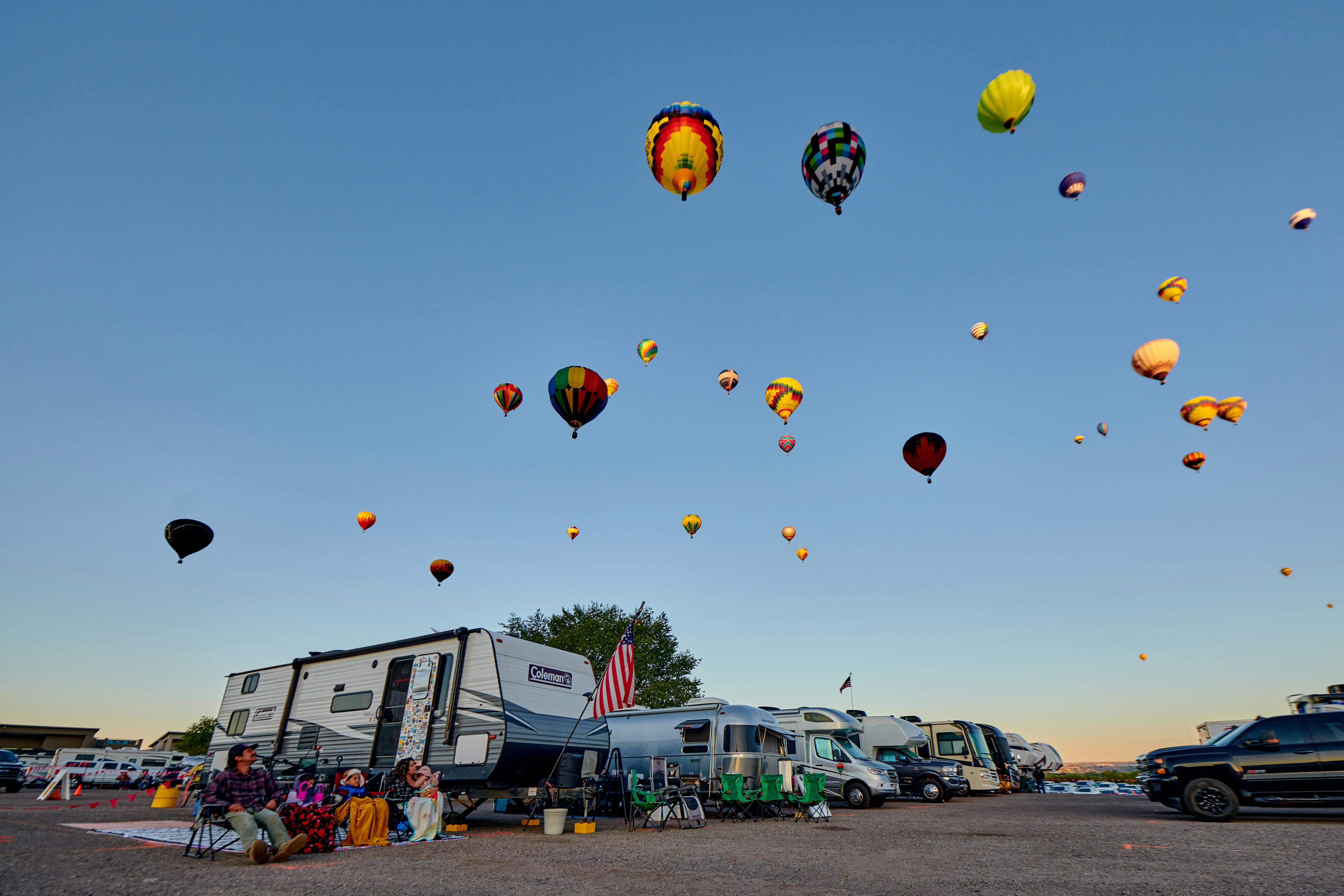 A coleman travel trailer parked in the RV lot of the Albuequerque Balloon Fiesta with Hot Air Balloons overhead. 