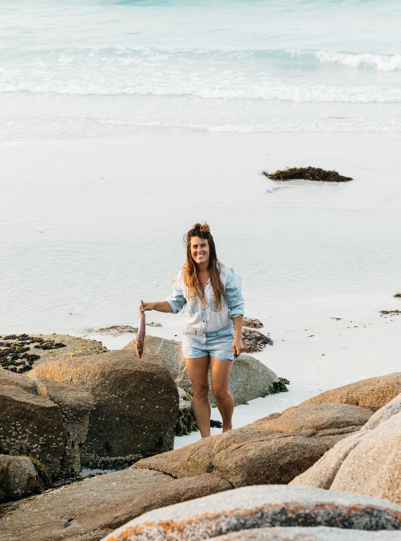 Sarah Glover on the beach, smiling at the camera, holding a fish.