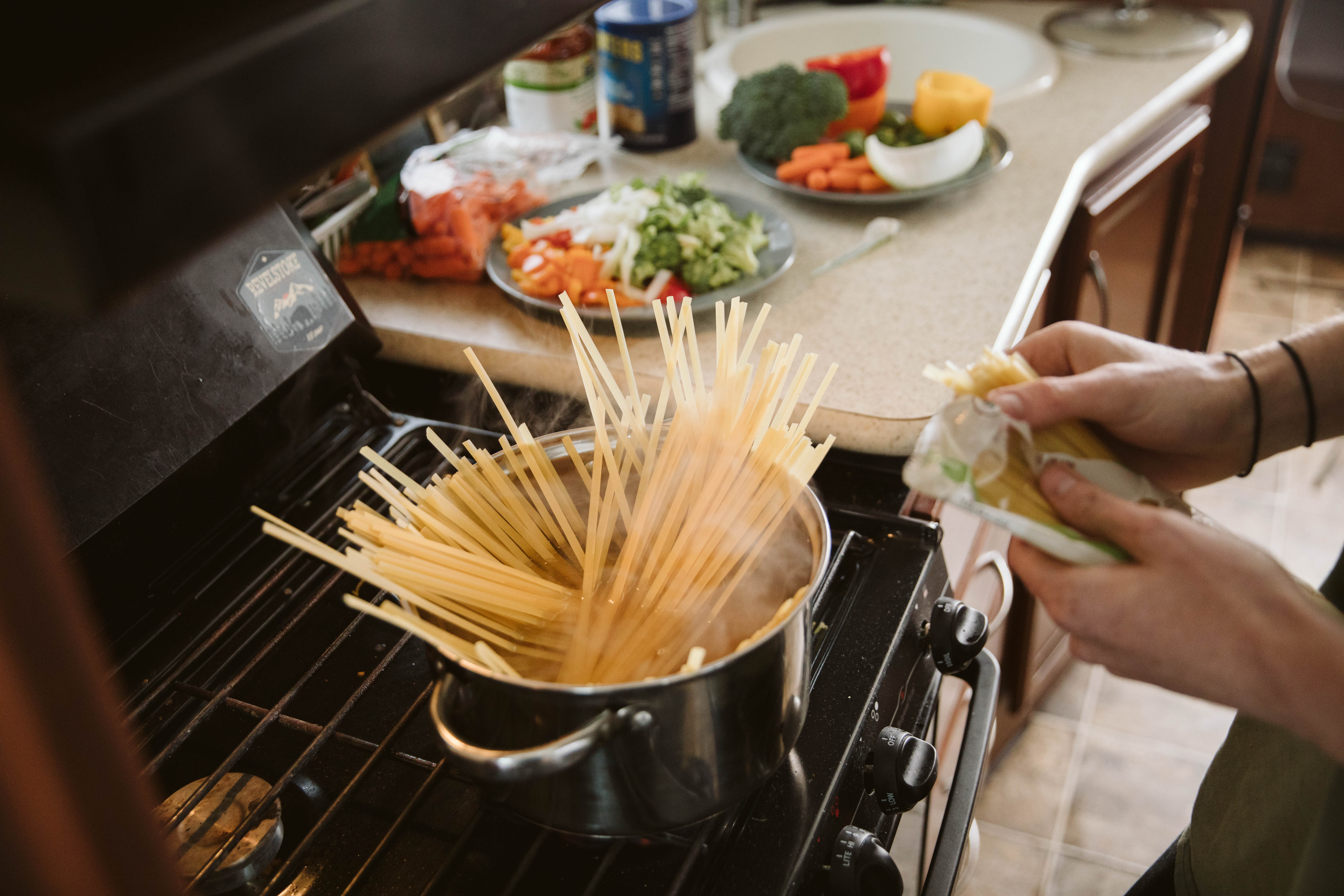 Close up of uncooked pasta in a pot of hot water with chopped vegetables in the background.
