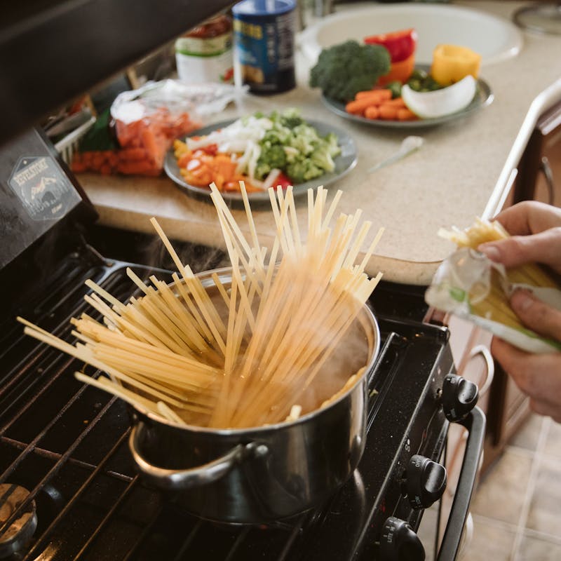 Close up of uncooked pasta in a pot of hot water with chopped vegetables in the background.
