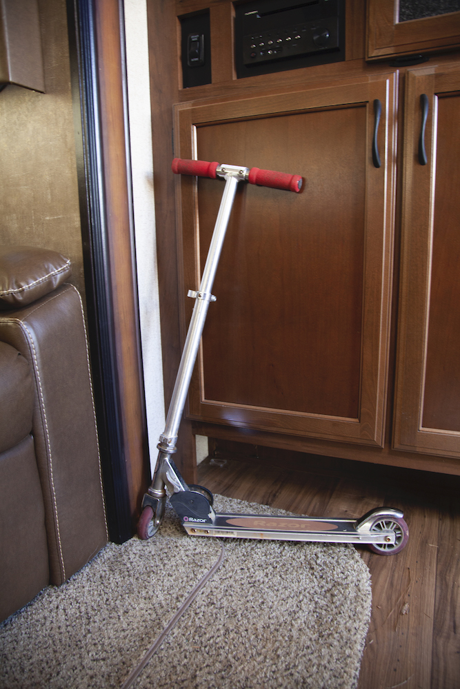 A slim foot-powered scooter leaning against a wall inside an RV. 