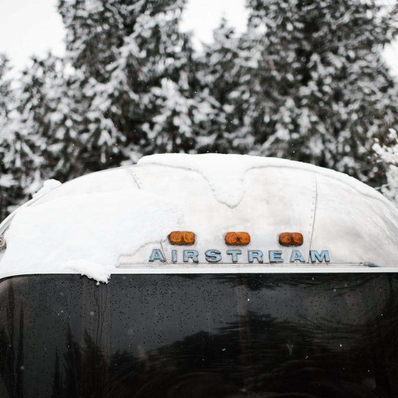 Front view of an Airstream RV, with snow on the roof, and snowy pine trees in the background.