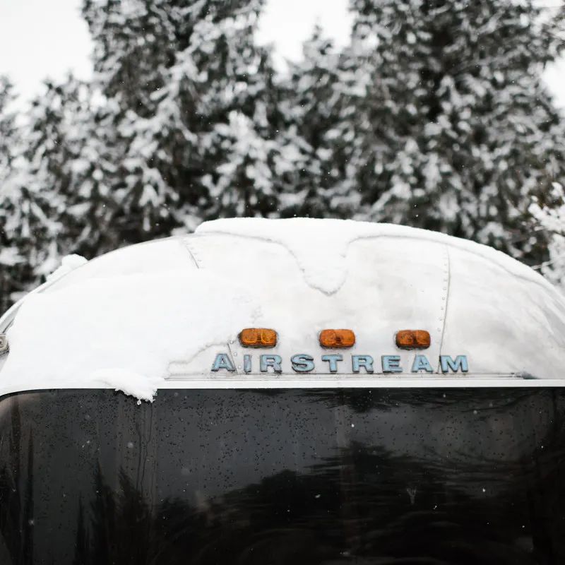 Front view of an Airstream RV, with snow on the roof, and snowy pine trees in the background.