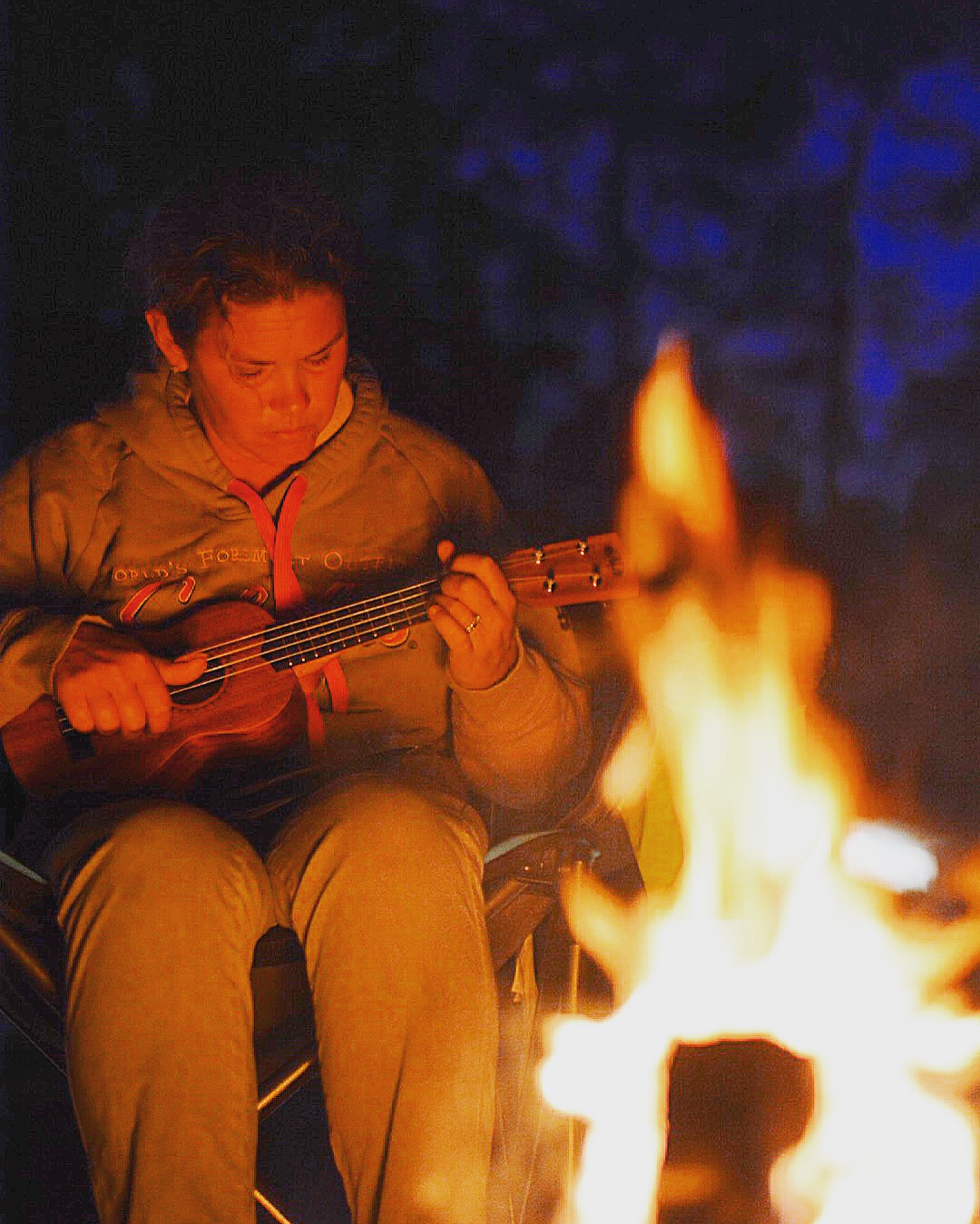 A woman playing a ukulele by the fire at night. 