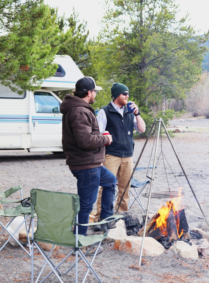 Two men stand around a camp fire, with a green camp chair and a Class C RV in the back.