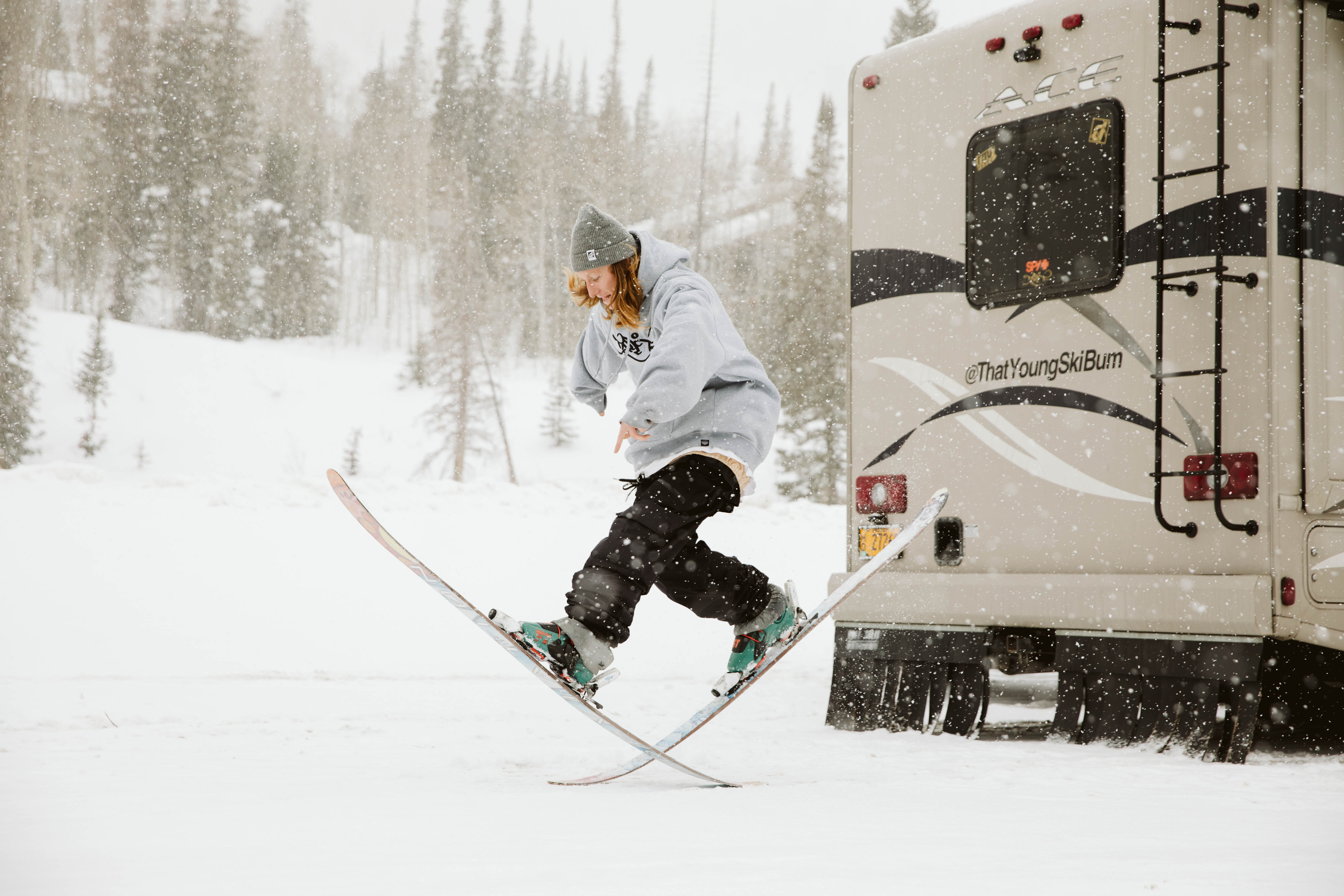 A guy on skis doing a trick jump at the bottom of a mountain next to an RV.