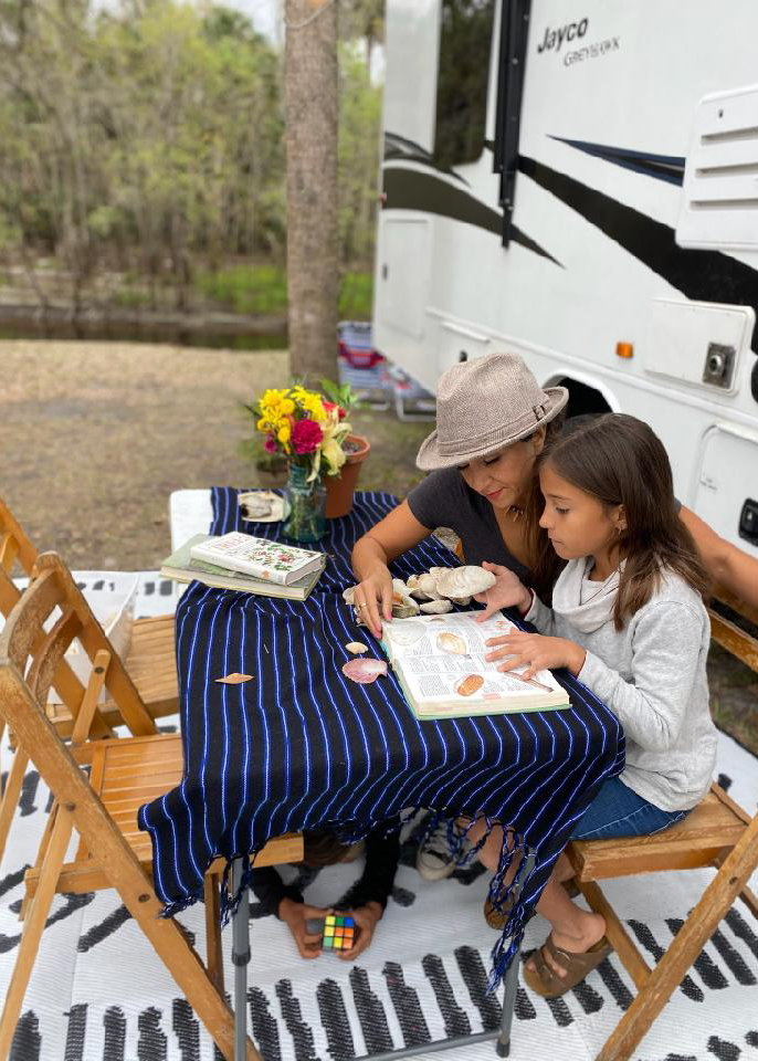 Sandra Peña and her daughter sit at a picnic table and identify shells in front of their Jayco Class C RV. 