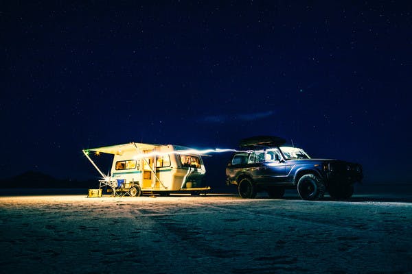 Dark photo with a blue car in front of a lit up trailer, with the awning out, door open, and chairs set up outside the trailer.