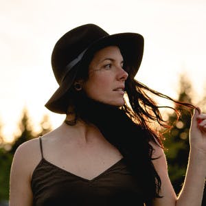 Aubrey Janelle, wearing a black hat and black shirt, holds hair and looks into the distance.