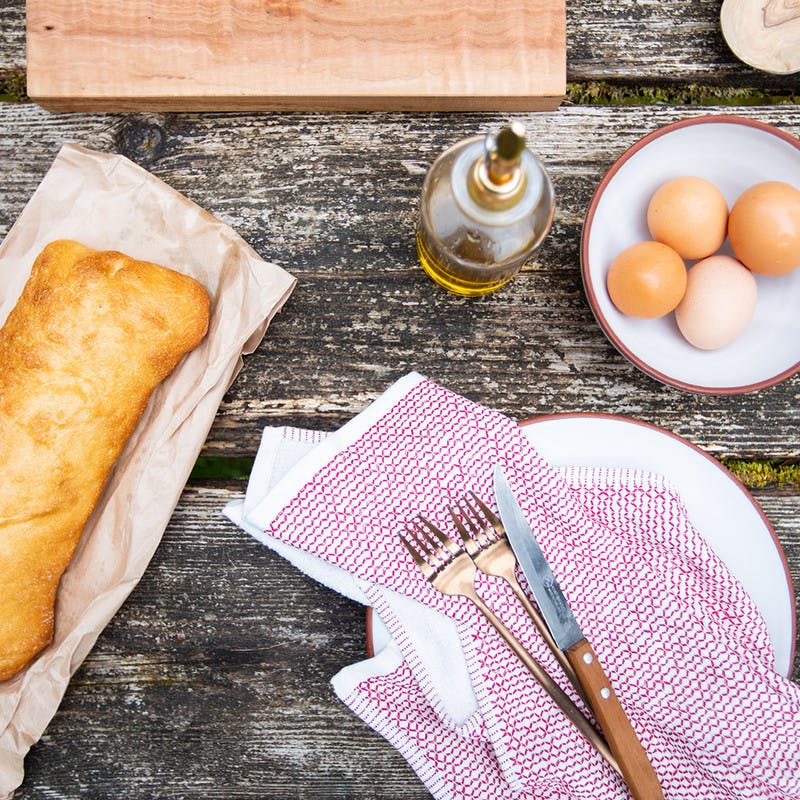 Artisan bread, eggs in a bowl, olive oil, and utensils with a napkin all sit on top of a picnic table to prepare for cooking a breakfast meal.