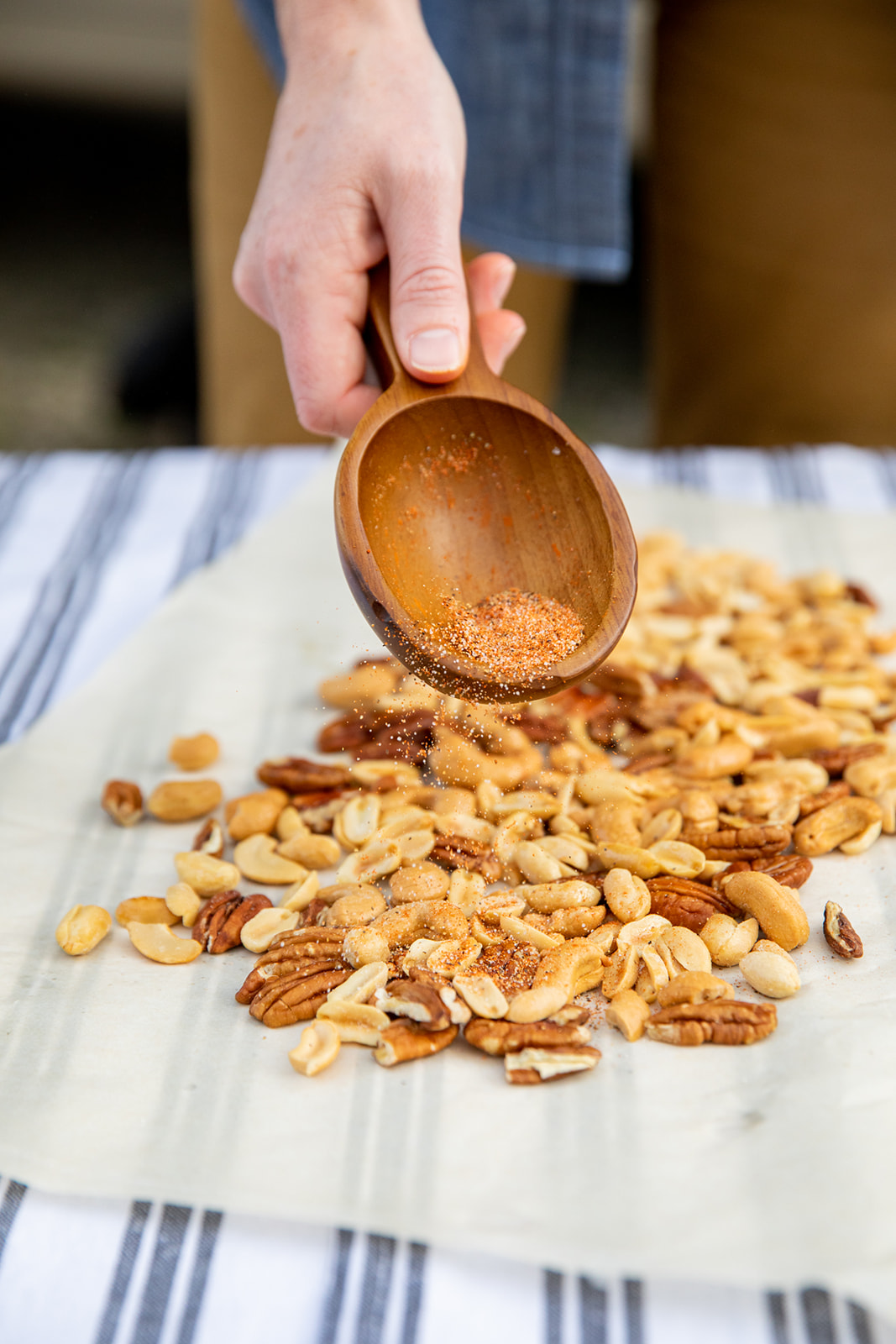 A hand holding a wooden spoon sprinkles spices over mixed nuts. 