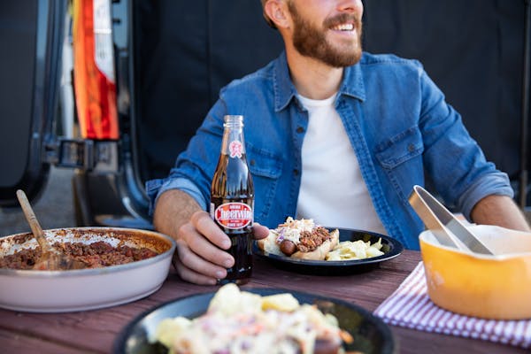 A man smiles holding a soda while sitting at a table and a Carolina Hot Dog plated in front of him.