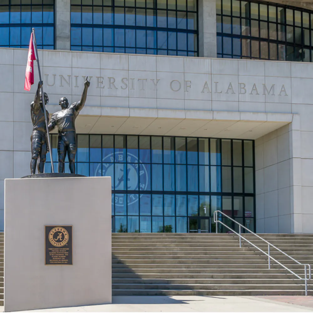 Brick front of Bryant–Denny Stadium at University of Alabama in Tuscaloosa, Alabama, with football player statues and American flag.