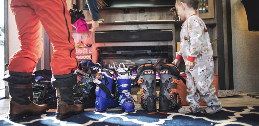 Ski boots are lined up by the fireplace inside an RV as a woman prepares to gear up for skiing.