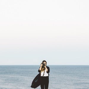 Image of Kat Parker looking through a camera lens with the ocean in the background. 
