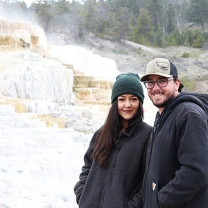 Juli and Jordan Cote standing in front of steaming geyser rocks, Juli with a green beanie on and Jordan with glasses and a baseball hat on.