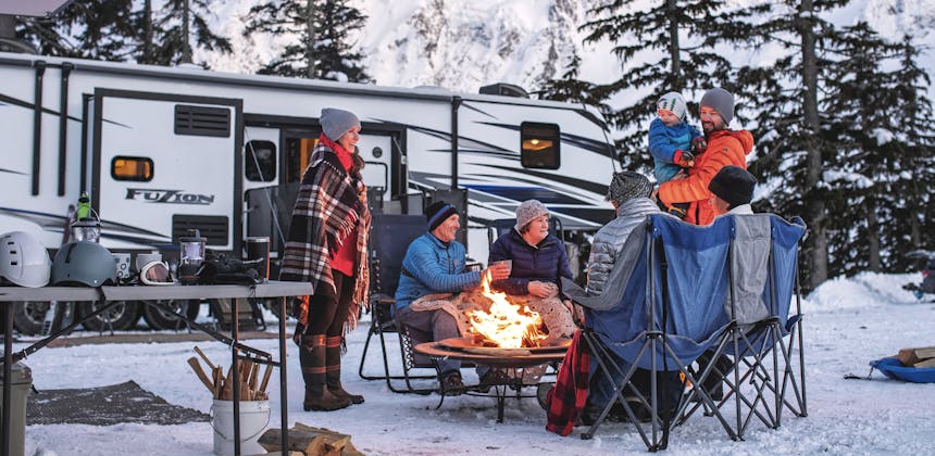 A family sits around a campfire outside of their RV, surrounded by snow.