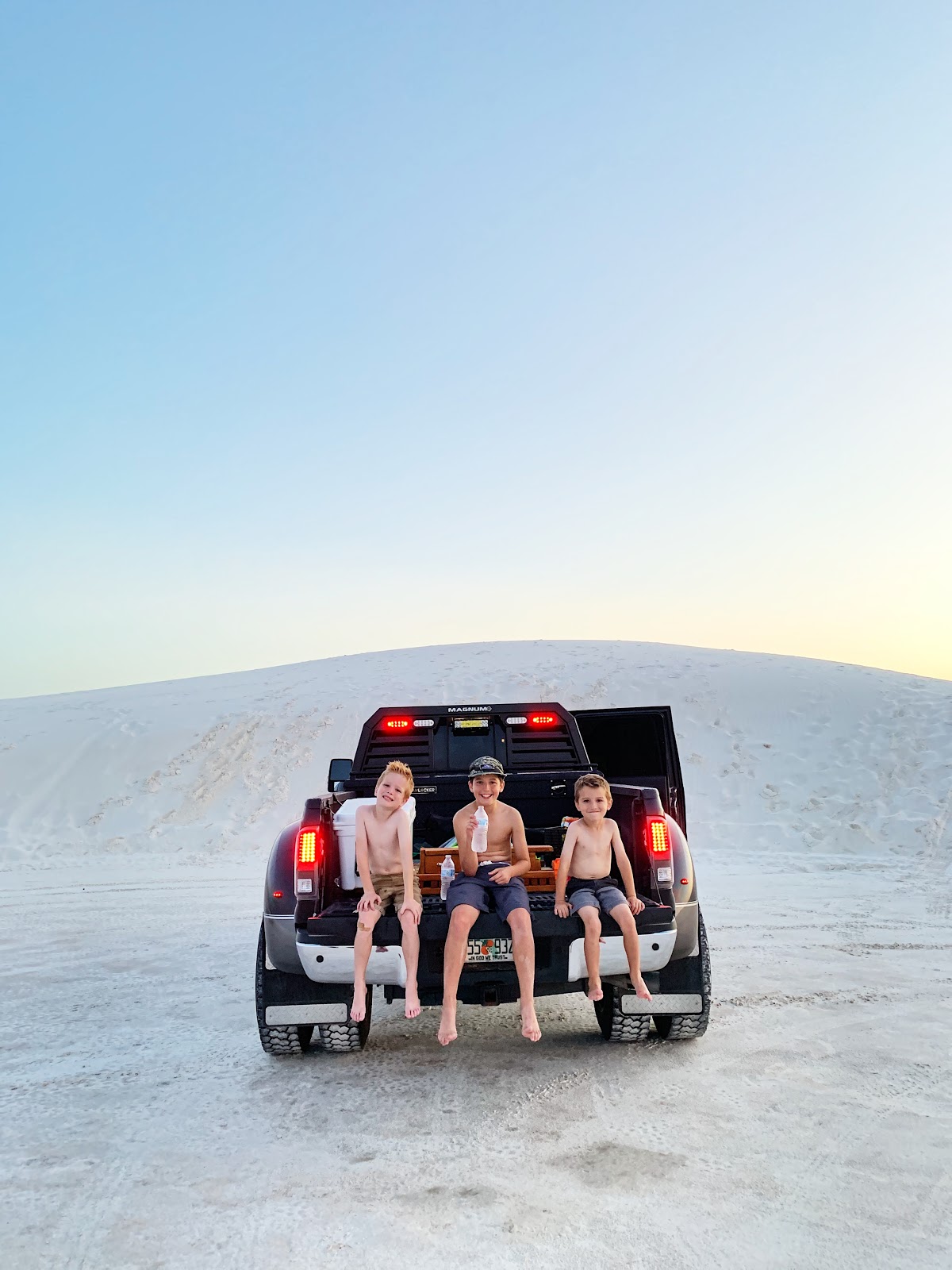 Three boys sitting on the open bed of a pickup truck while it's parked.